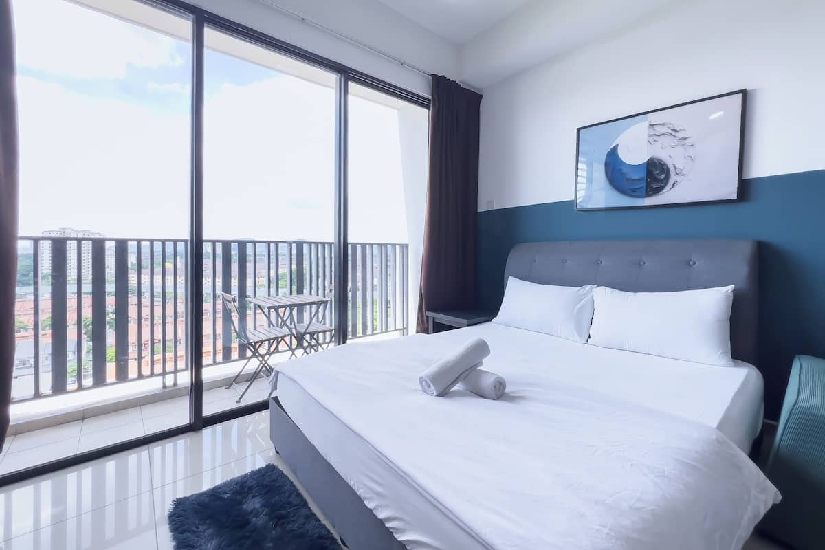 Une chambre d’hôtel dotée d’un grand lit, offrant une vue sur la ville grâce à de larges fenêtres du sol au plafond, et disposant d’un balcon avec une table et des chaises.