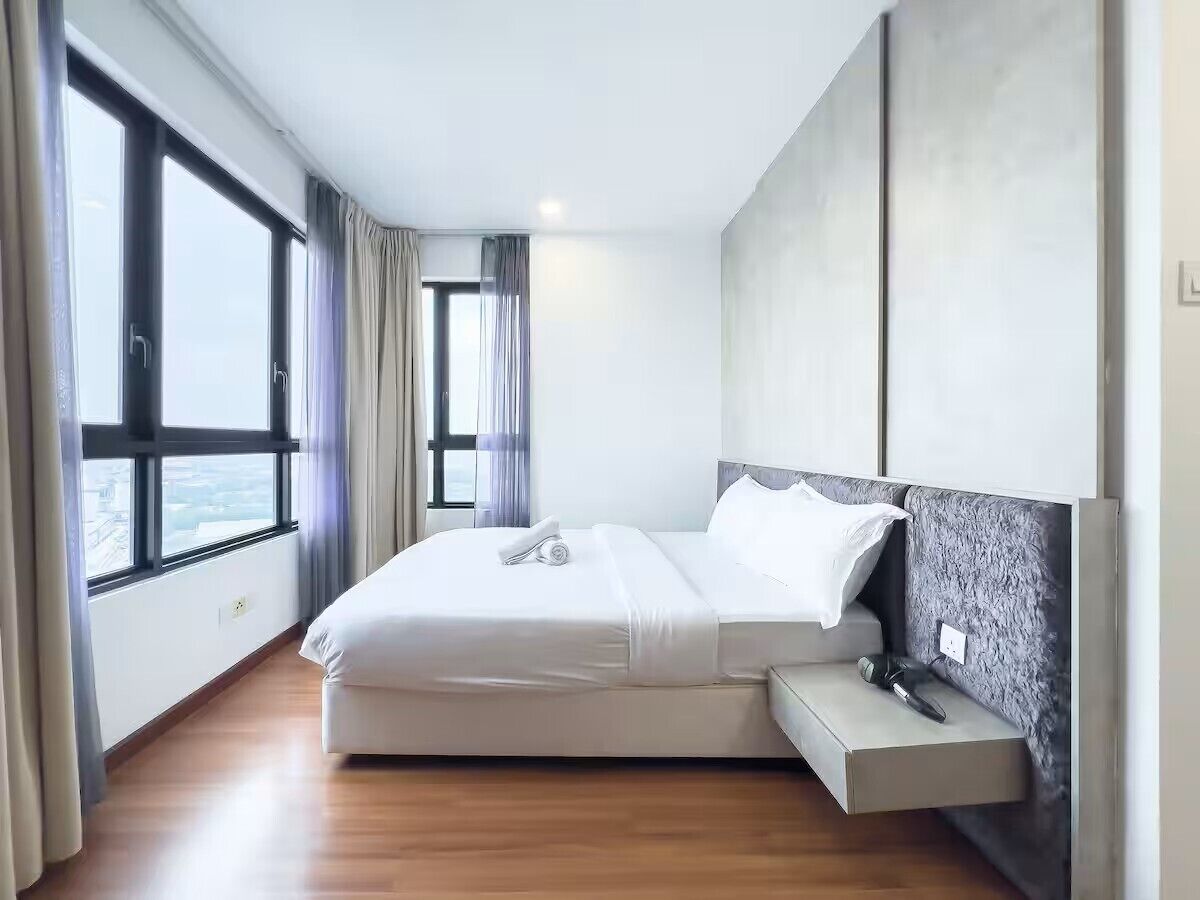 A modern hotel room with a large bed, a bedside table, and a view of the city through large windows.
