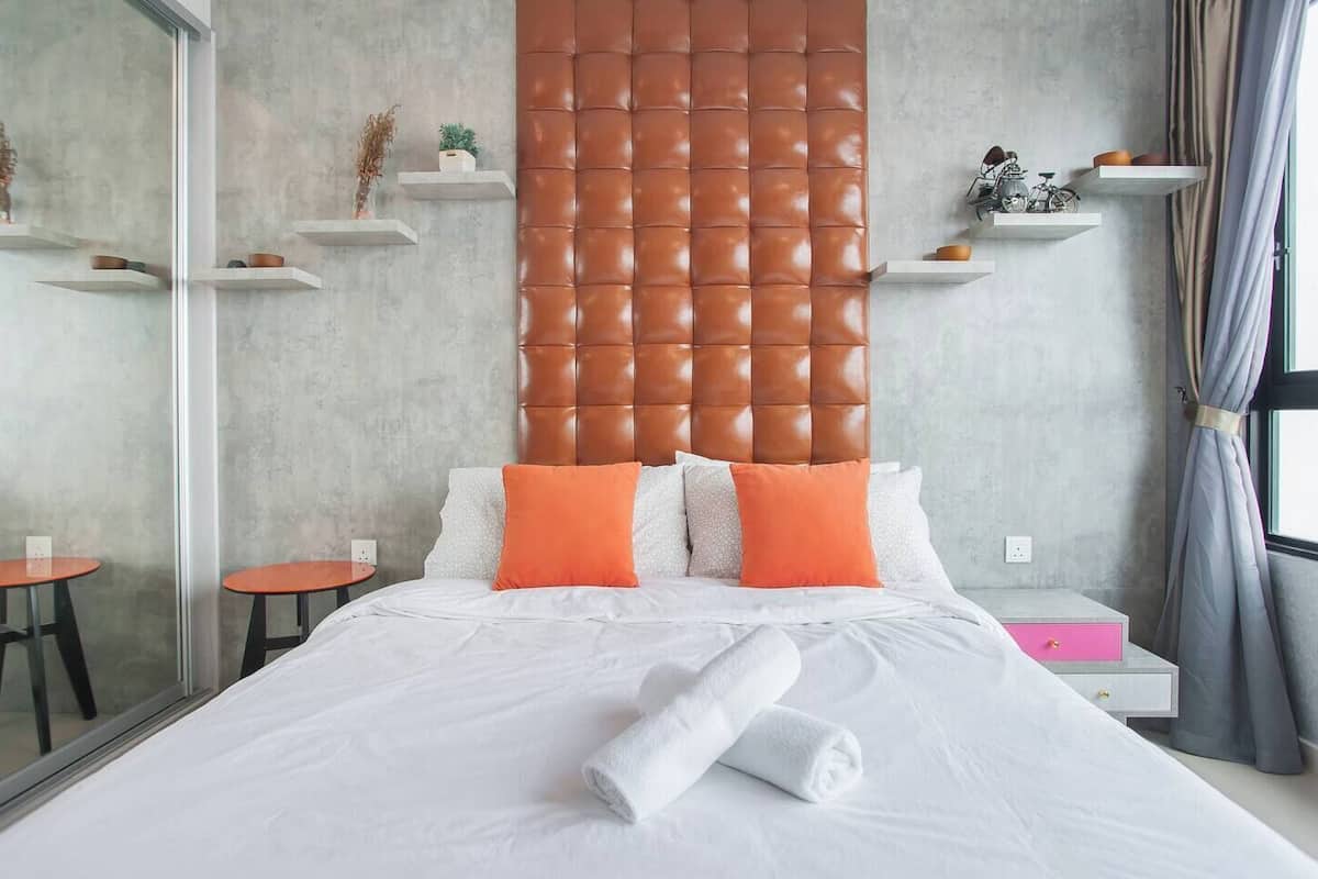 Une chambre d’hôtel moderne avec un grand lit, des oreillers orange, une tête de lit capitonnée et une cloison en verre.