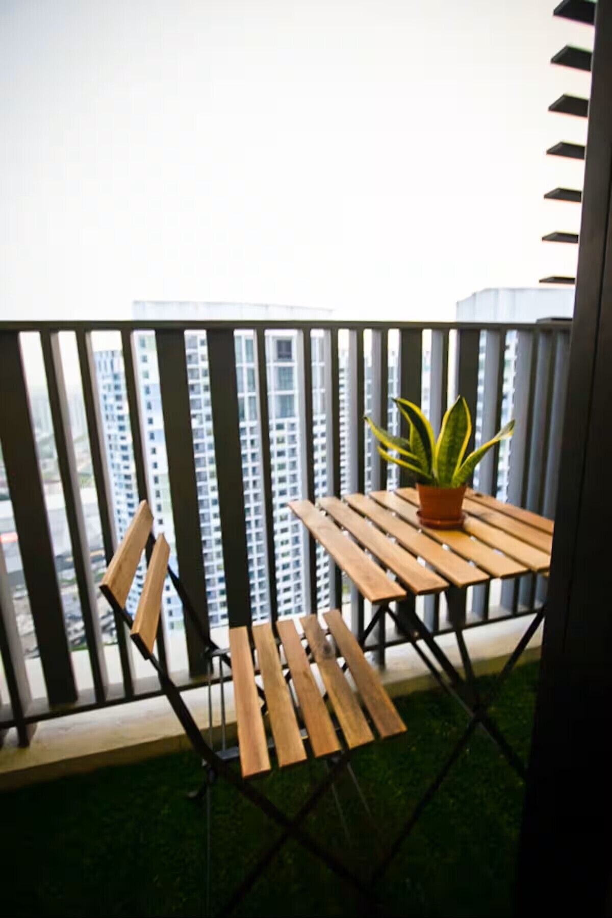A balcony with a wooden table and chairs, a potted plant, and a view of high-rise buildings.