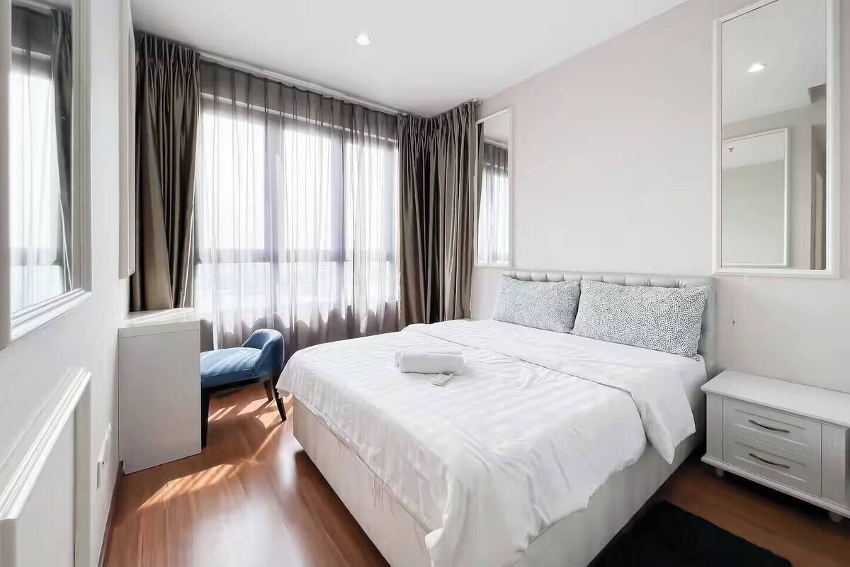 Moderni hotellihuone, jossa on suuri sänky, yöpöytä, tuoli ja ikkuna, jossa on verhot.