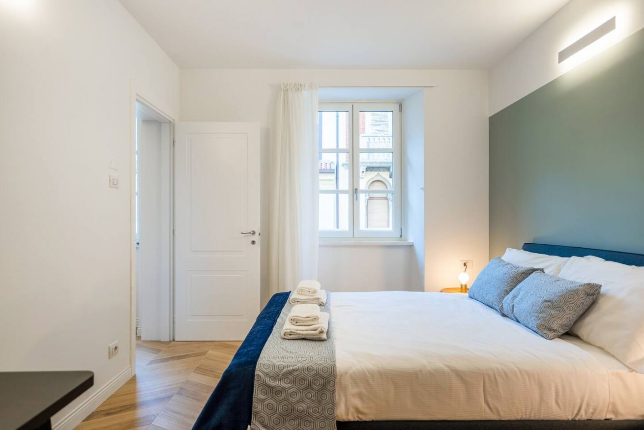 Un dormitorio con cama, mesita de noche, ventana con cortinas y mesita auxiliar con lámpara.