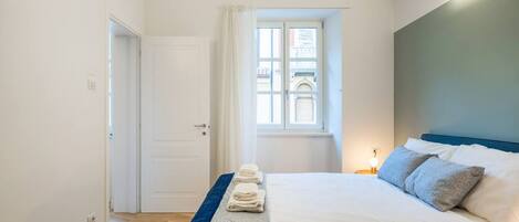 Un dormitorio con cama, mesita de noche, ventana con cortinas y mesita auxiliar con lámpara.