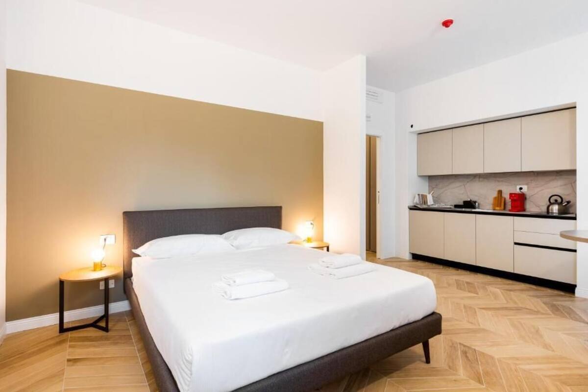 A hotel room with a bed, bedside lamps, a kitchen area, and a herringbone-patterned floor.