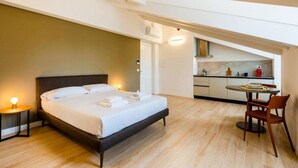 A bedroom with a large bed, a bedside table with a lamp, and a view into a kitchen area with a dining table and chairs. - JOIVY Premium Apartments Trieste Mazzini (Trieste)