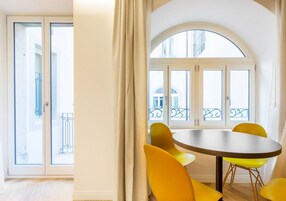 Una stanza con un tavolo rotondo e sedie gialle, un'ampia finestra ad arco e una porta scorrevole in vetro che dà su un balcone esterno.