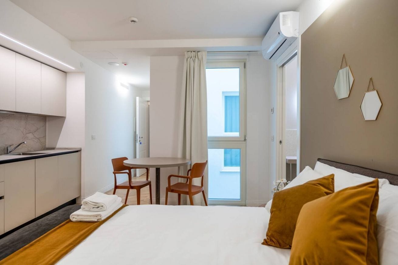 A hotel room with a bed, a small table with two chairs, a kitchen area, and a balcony with a view.