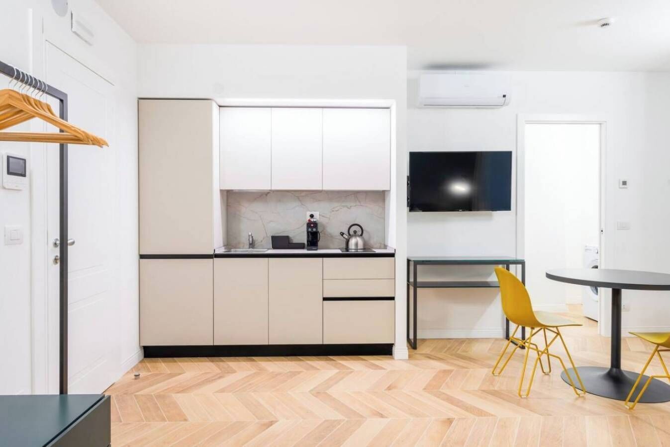 A modern kitchen with white cabinets, a black countertop, and a kettle. There is a dining area with a table and yellow chairs.