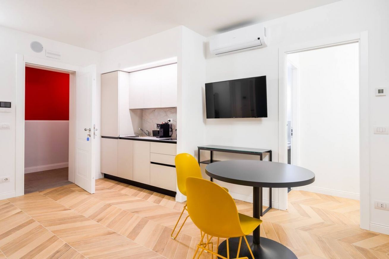 A modern kitchen with white cabinets, a red accent wall, and a dining area with a round table and yellow chairs.