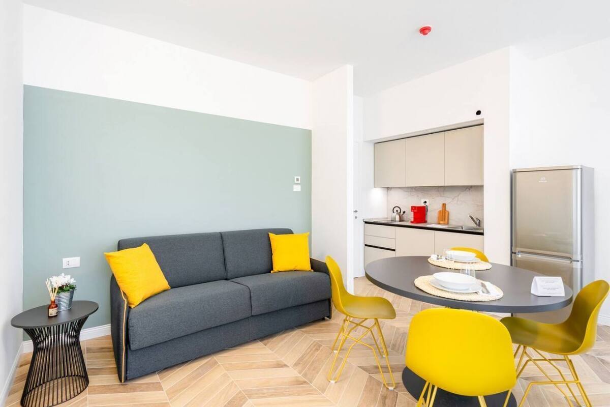 A modern living room with a grey sofa, yellow cushions, a round dining table with yellow chairs, and a kitchen area with cabinets and appliances.