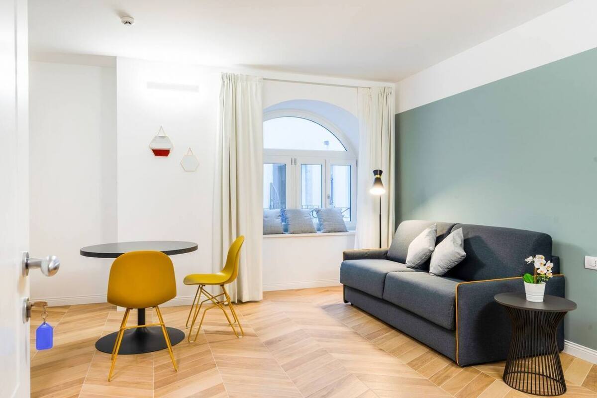 A modern living room with a sofa, a small round table, and two chairs. There is a window with curtains and a view of the outside.