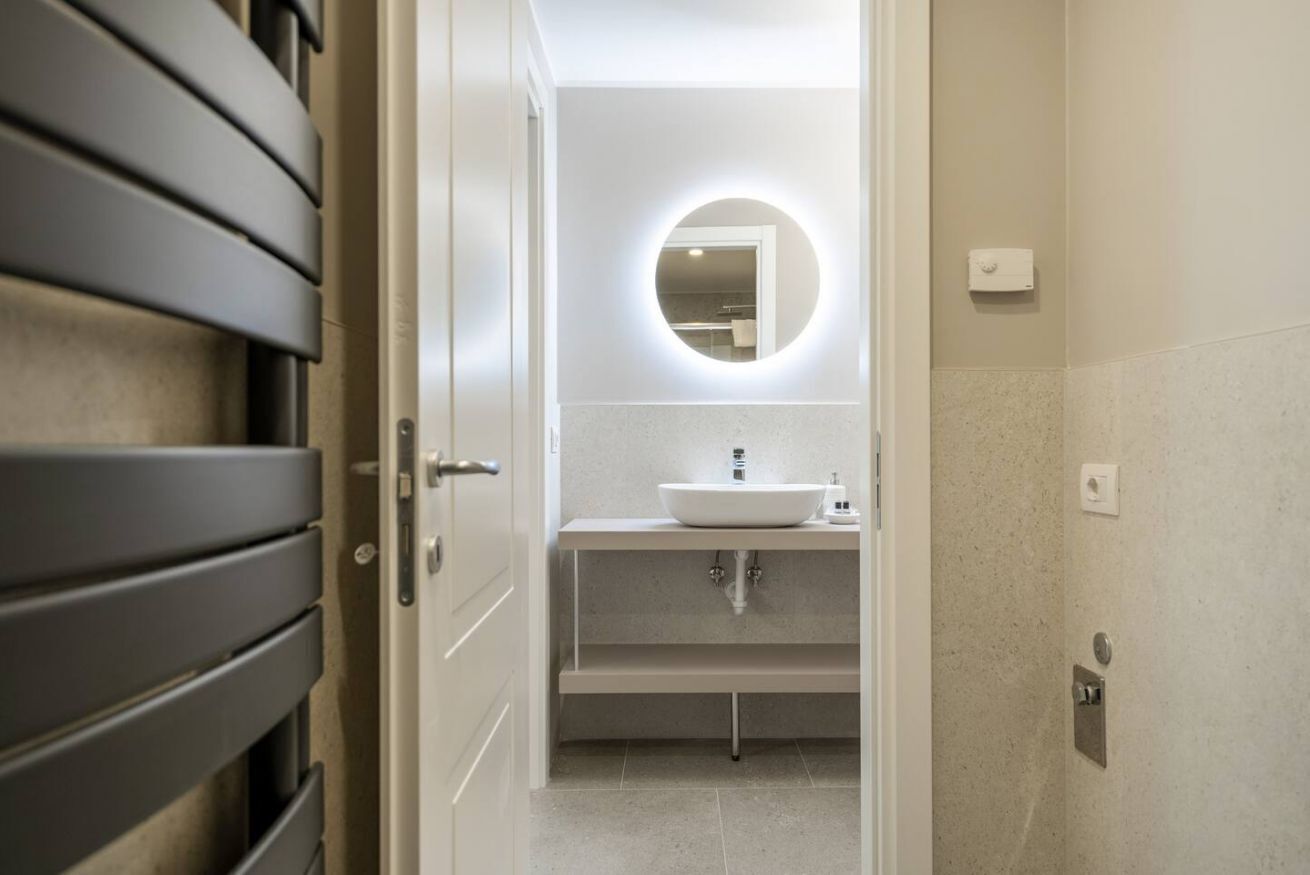 Une salle de bain moderne avec un lavabo blanc, un miroir rond et une porte dotée de poignées horizontales.