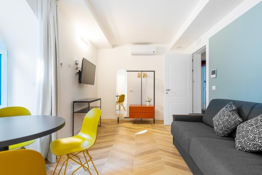Un salon moderne avec un canapé gris, des fauteuils jaunes, une petite table et un téléviseur fixé au mur.