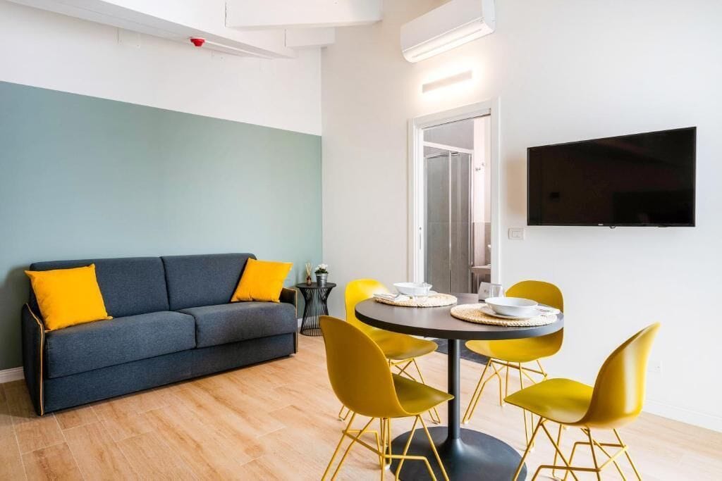 A modern living room with a sofa, yellow chairs, a dining table, and a flat-screen TV mounted on the wall.