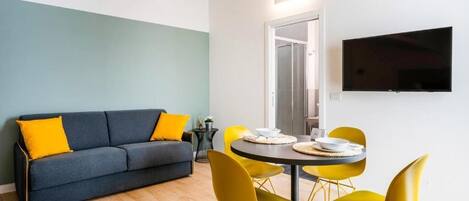 A modern living room with a sofa, yellow chairs, a dining table, and a flat-screen TV mounted on the wall.