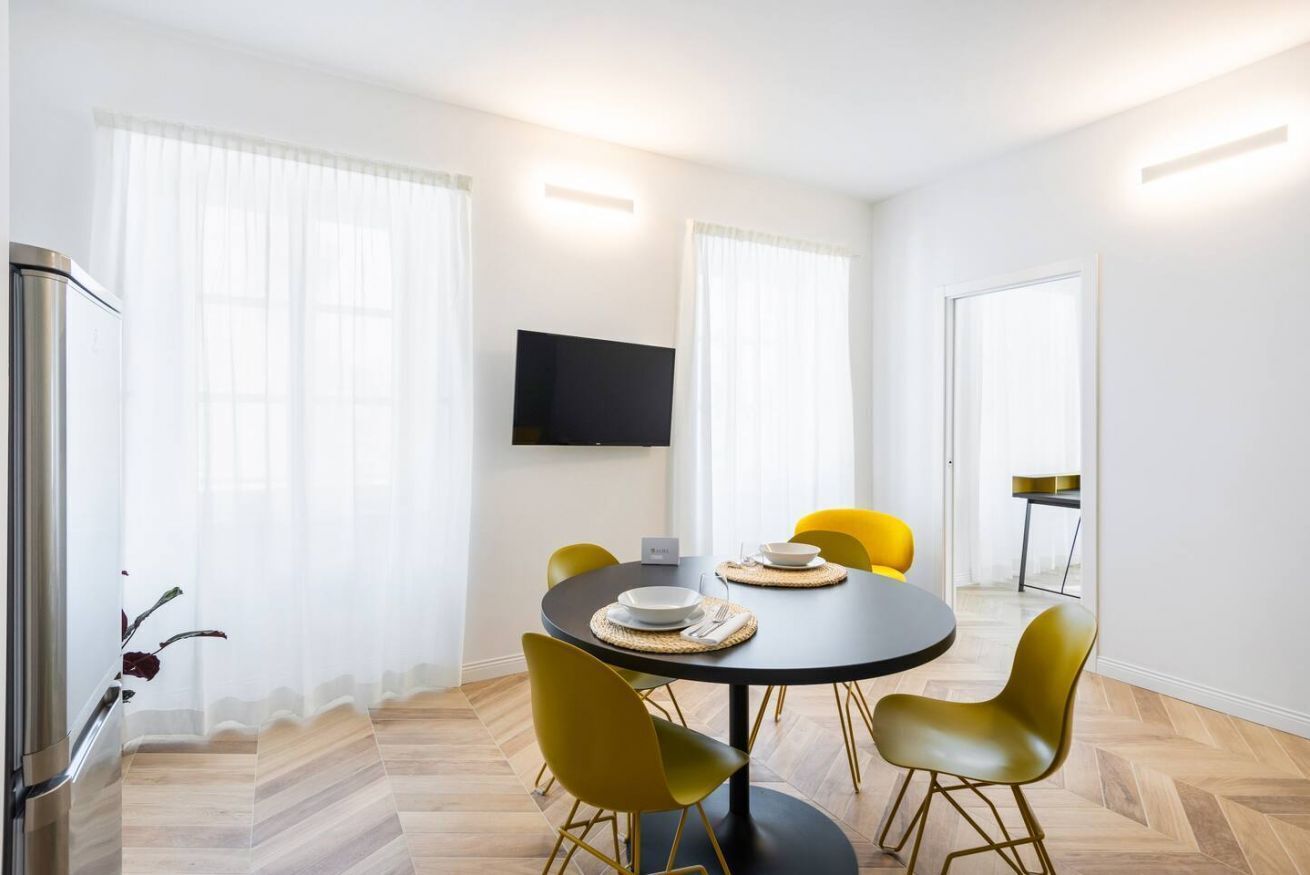 A modern dining area with a round table, yellow chairs, and a flat-screen TV mounted on the wall.