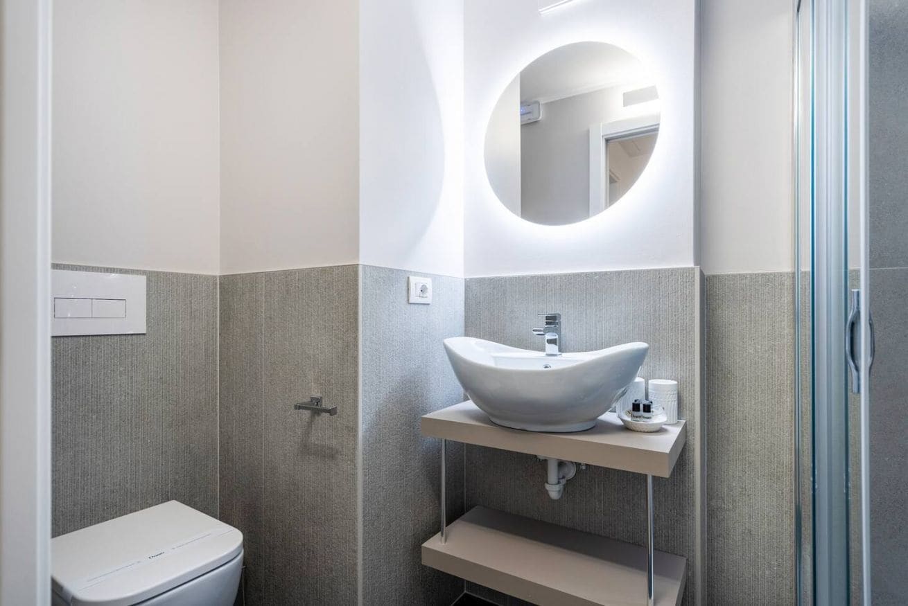 A modern bathroom with a white toilet, a vessel sink, a round mirror, and a shower area.