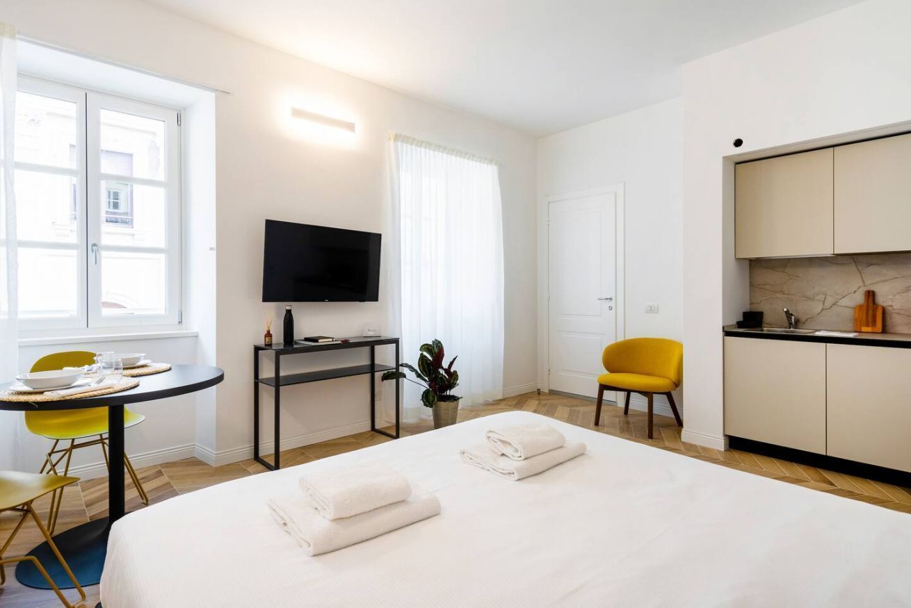 Habitación de hotel con cama, escritorio con televisor, una silla, una mesita con planta y un área de cocina.