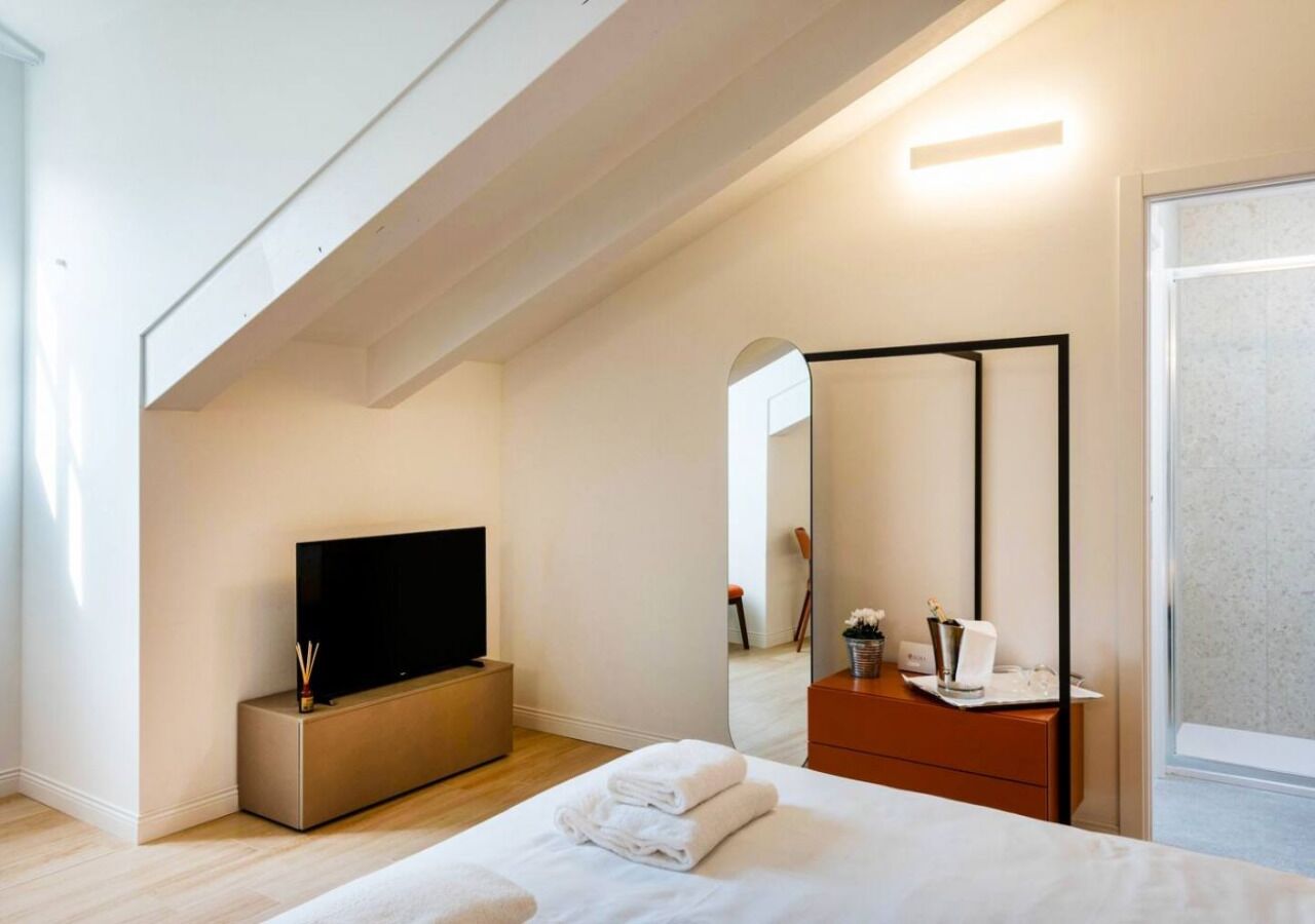 Una habitación de hotel moderna con una televisión de pantalla plana, una cama con toallas blancas ordenadas, un tocador de madera y un espejo grande.