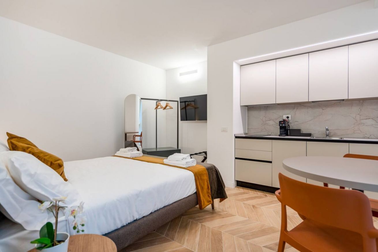 Habitación de hotel con cama, mesita, silla, espejo, armario y kitchenette.