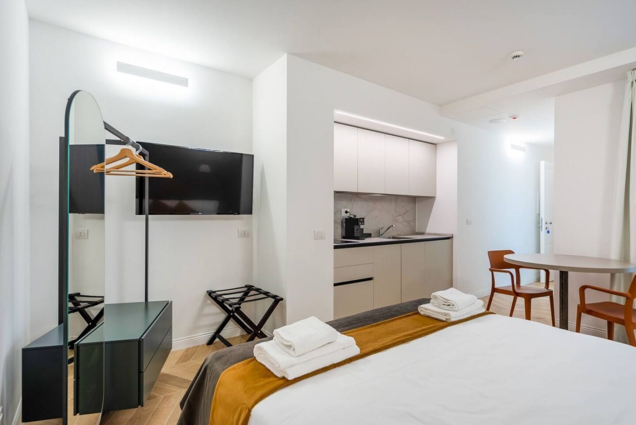 Ein modernes Hotelzimmer mit Bett, Schreibtisch, Stuhl, Fernseher und einer Küchenzeile.
