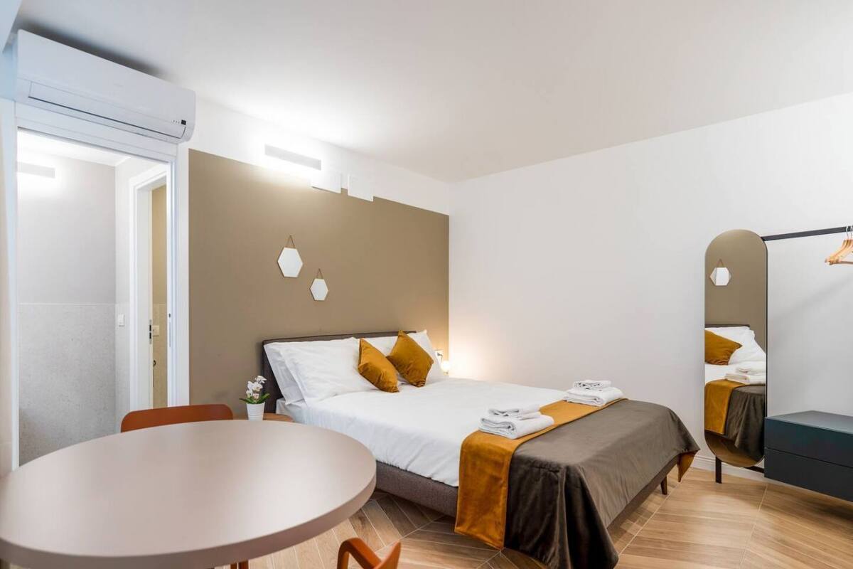 A hotel room with a bed, a table, and a chair. There is a mirror on the wall and a door leading to another room.