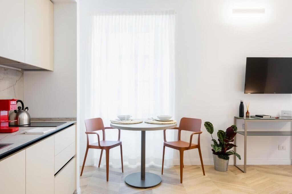 A modern kitchen with a dining area, including a round table with chairs, a coffee machine, and a TV mounted on the wall.