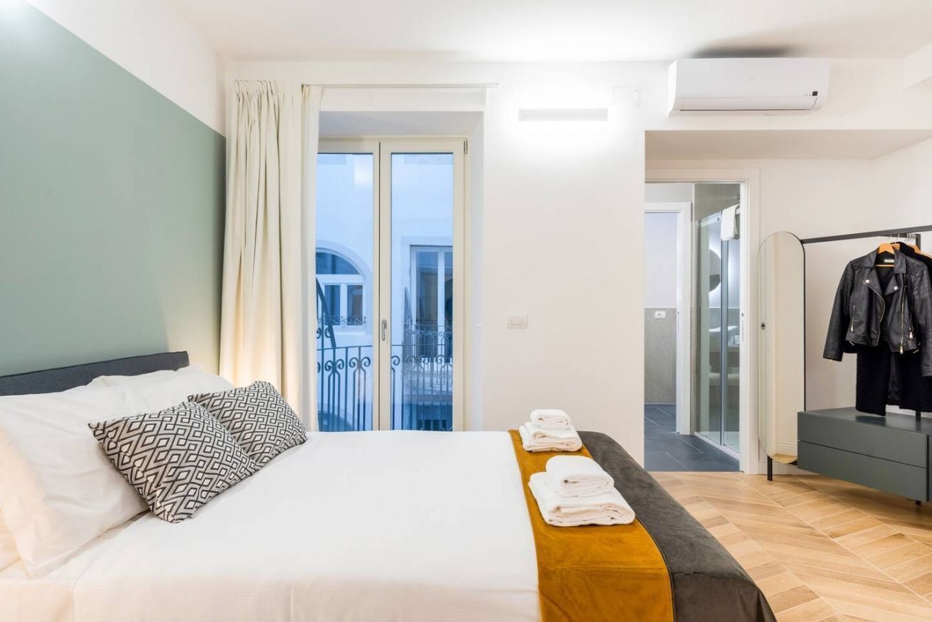 A hotel room with a bed, a wooden bedside table, a wardrobe, and a balcony with a view.