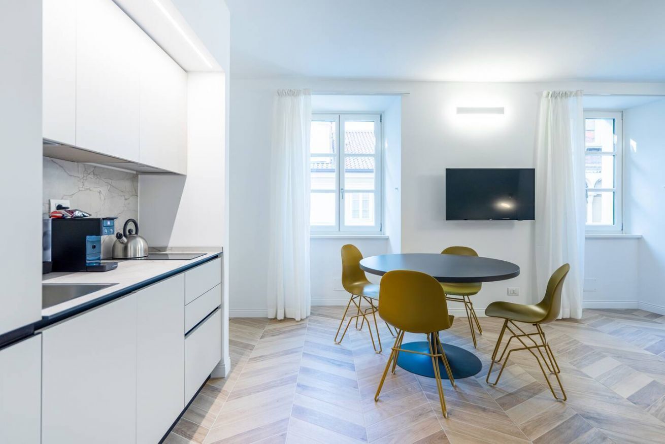 Une cuisine moderne avec des meubles blancs, un plan de travail noir et une bouilloire. On y trouve un coin repas avec une table ronde et quatre chaises. Un téléviseur à écran plat est fixé au mur, au-dessus de la table.