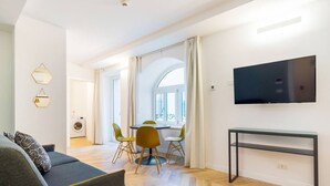 A modern living room with a sofa, a dining table with chairs, a TV mounted on the wall, and a herringbone-patterned floor. - JOIVY Premium Apartments Trieste Mazzini (Trieste)