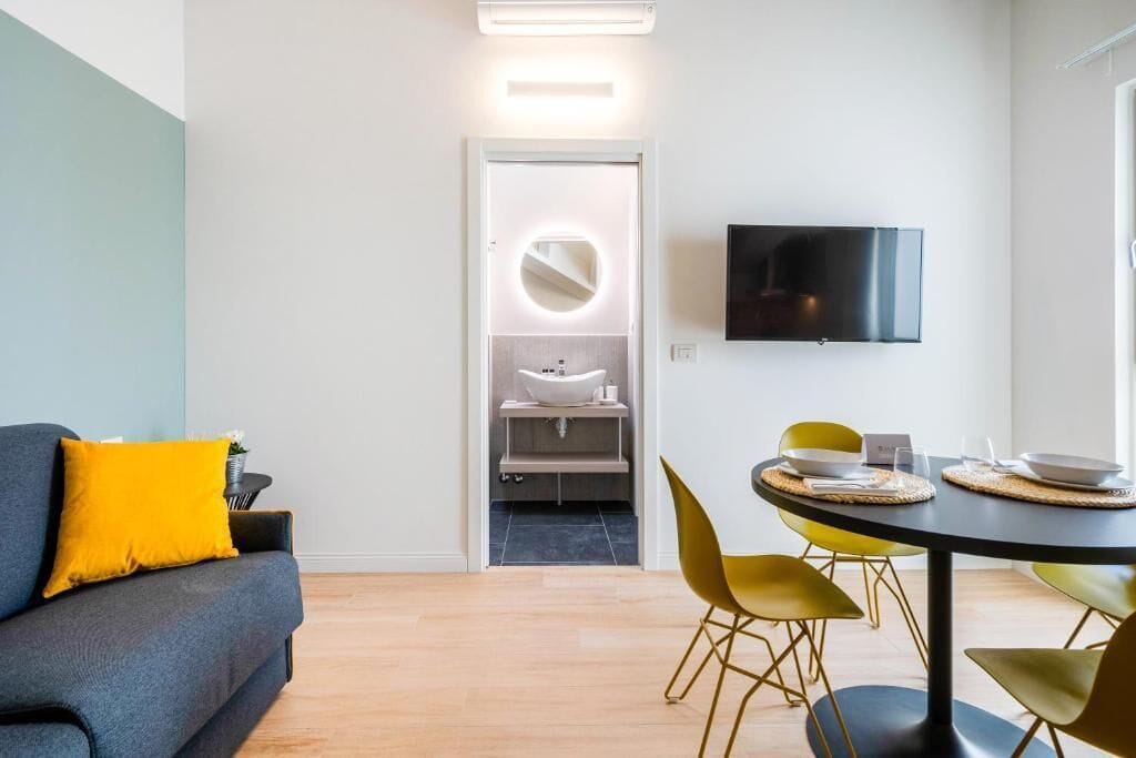 Ein modernes Wohnzimmer mit Sofa, einem Esstisch mit Stühlen, einem an der Wand befestigten Fernseher und einem Badezimmer mit Waschbecken und Spiegel.