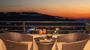 Ein Esstisch mit Korbstühlen, eine Schüssel mit Essen und zwei Gläser Orangensaft auf einem Balkon mit Blick auf eine Stadtlandschaft in der Abenddämmerung.