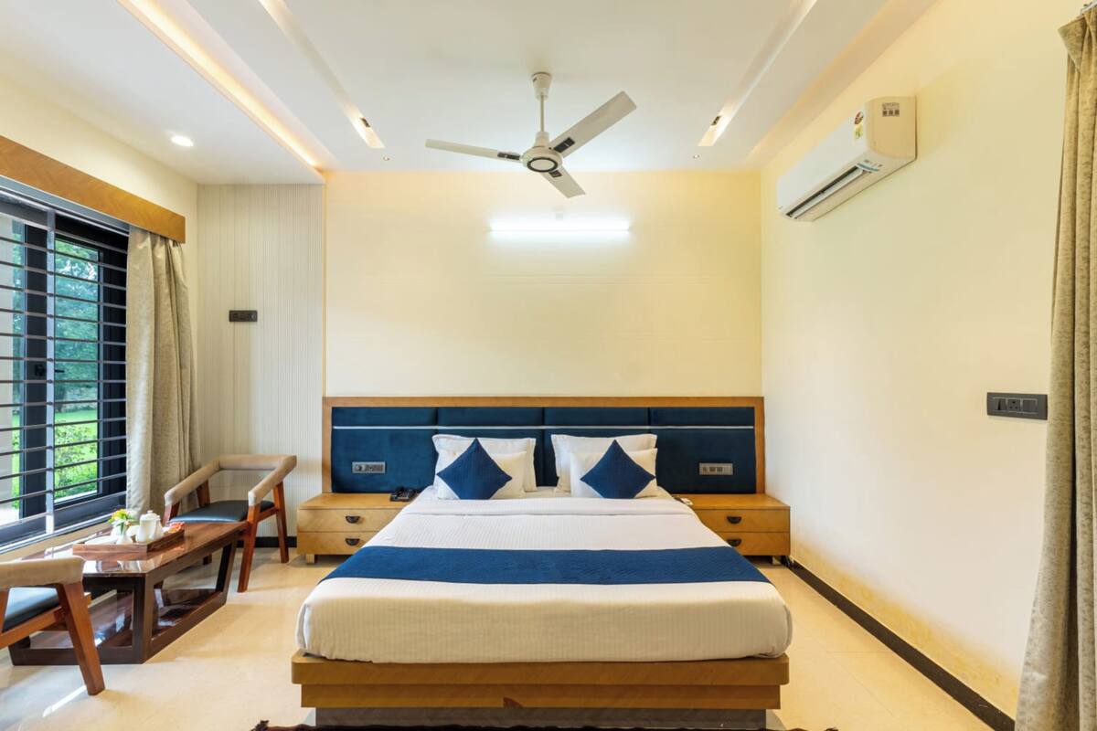A hotel room with a bed, bedside tables, a desk, a chair, a ceiling fan, and an air conditioning unit.