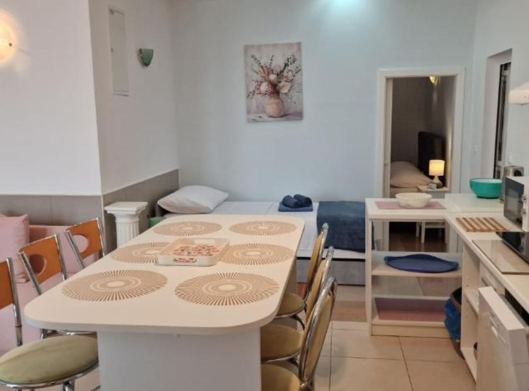A compact living space with a dining table, chairs, a bed, a small kitchenette, and a painting on the wall.