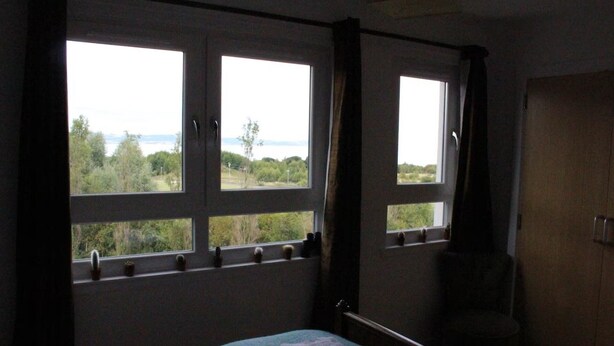 A bedroom with a bed, a chair, and two windows offering a view of greenery and a body of water. - 2-bed Seaside Apartment - Sleeps 5 & Free Parking (Edinburgh)