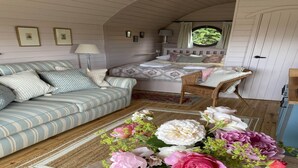 A cozy room with a striped sofa, a bed with a floral bedspread, a wicker chair, and a vase of flowers on a wooden floor. - Cabins in Oxfordshire with Hot Tub & Meadow Views (Banbury)