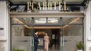 The entrance of PCAHOTEL with a glass door, a signboard, and a potted plant outside.