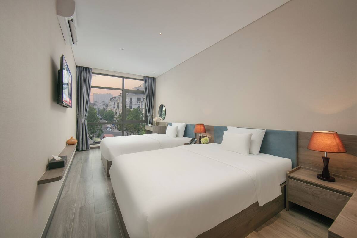 Una habitación de hotel con dos camas, una mesita de noche con lámpara, una pequeña mesa con un florero y una ventana con cortinas.