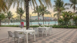 An outdoor seating area with tables and chairs, palm trees, and a view of a beach and hotel in the background.