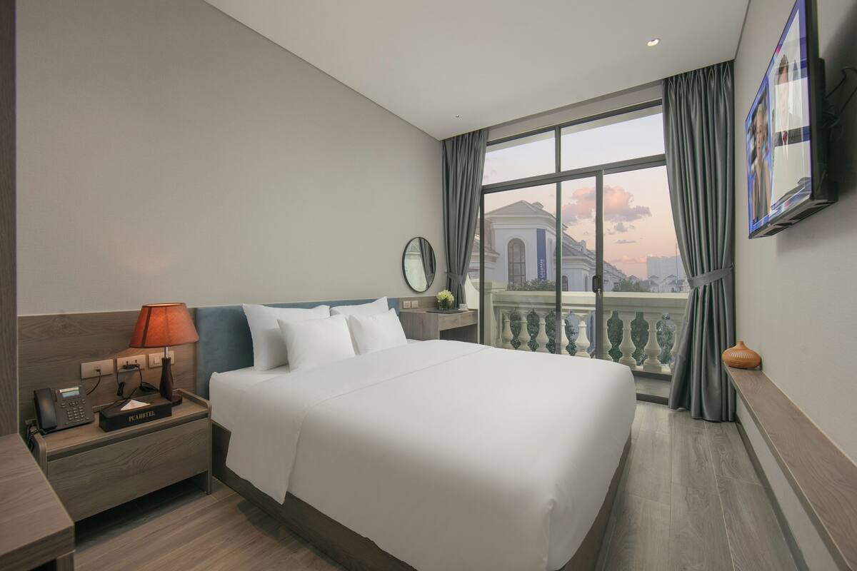 Habitación de hotel con una cama grande, una mesita de noche con lámpara, un teléfono y vistas a un edificio a través de una ventana con cortinas.