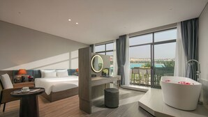 Une chambre d’hôtel moderne dotée d’un grand lit, d’un bureau, d’un miroir rond, d’une baignoire remplie de pétales de rose et d’un balcon offrant une vue sur le paysage urbain.