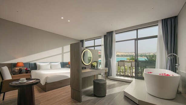 A modern hotel room with a large bed, a desk, a round mirror, a bathtub with rose petals, and a balcony with a view of a cityscape.