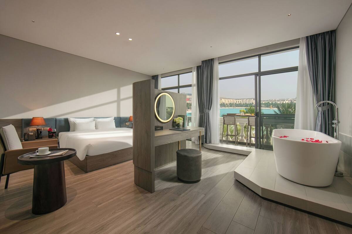 Una habitación de hotel moderna con una cama grande, un escritorio, un espejo redondo, una bañera con pétalos de rosa y un balcón con vistas a un paisaje urbano.