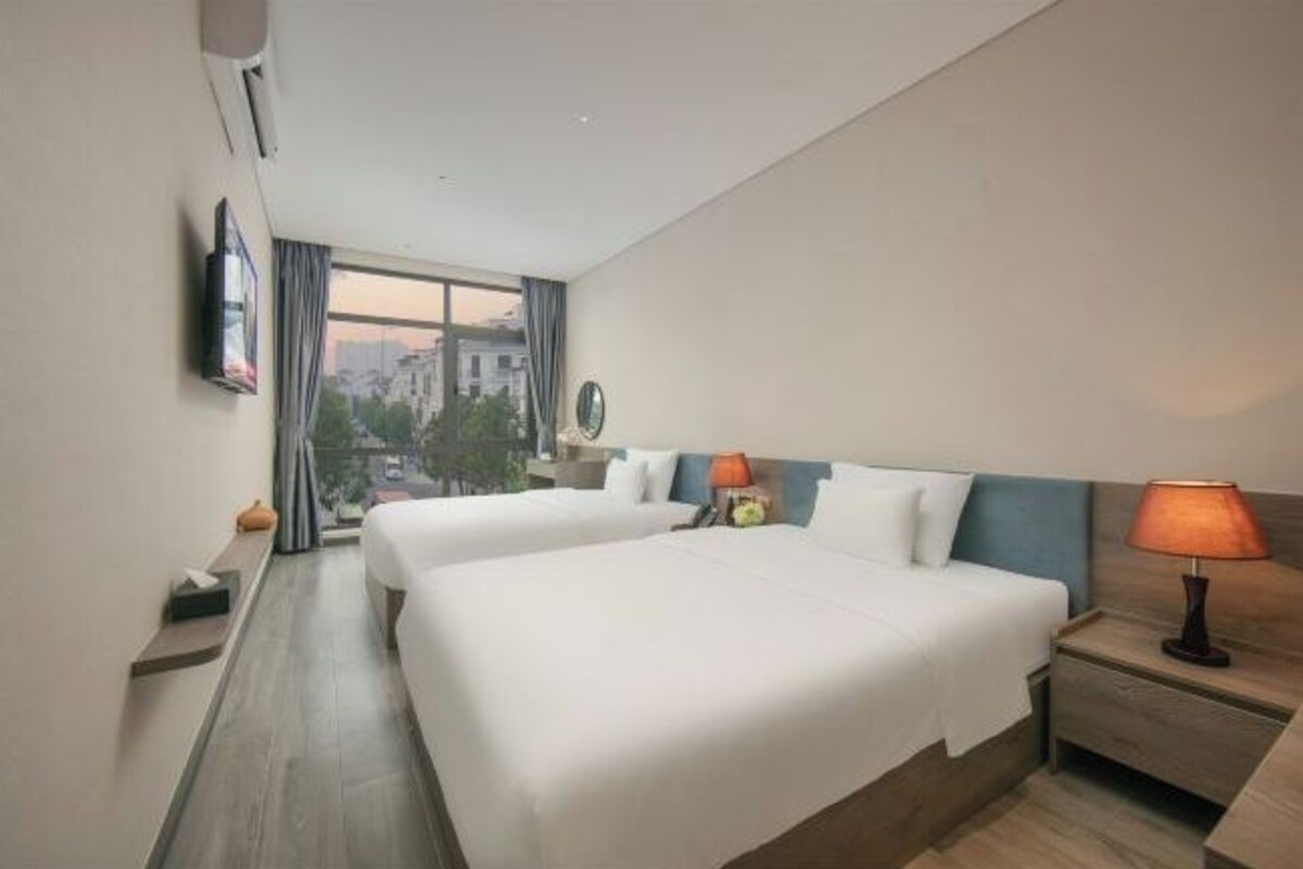 Een hotelkamer met twee bedden, een nachtkastje, een lamp, een aan de muur bevestigde televisie en uitzicht op de stad door grote ramen.