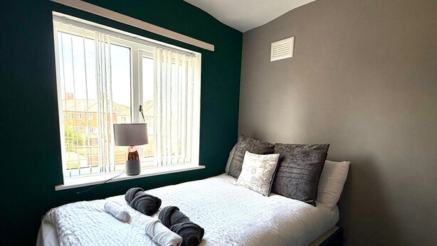 Cama bem arrumada com roupa de cama branca e cinza, um abajur no parapeito da janela e vista para prédios lá fora.