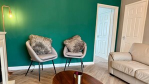 Sala de estar com paredes verde-água, dois poltronas com almofadas de pele, uma mesa de centro de madeira, um sofá bege e uma porta branca.