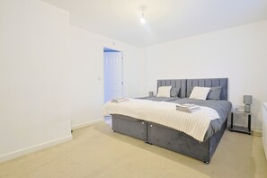Ein modernes Schlafzimmer mit einem grau gepolsterten Bett, weißer Bettwäsche und einem Nachttisch mit einer Lampe.