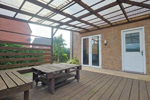 Ein überdachter Patio-Bereich mit einem Holztisch und Bänken, einer Backsteinmauer und einer Tür mit Glasfenstern.