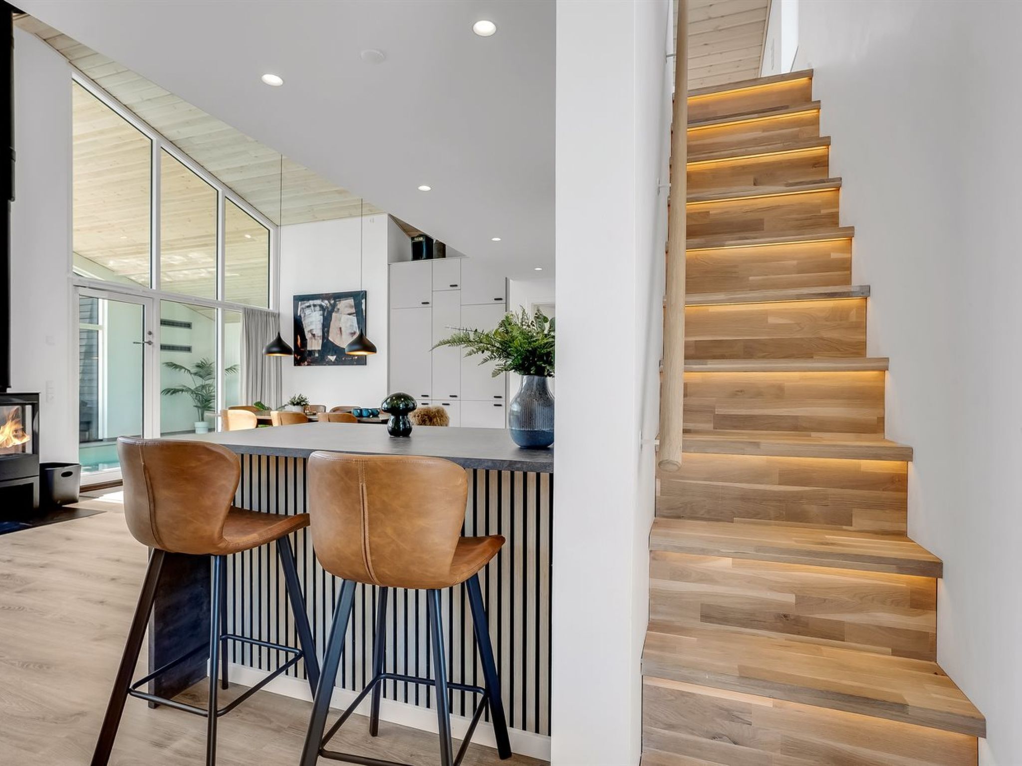 A modern kitchen with a central island, bar stools, and a staircase with wooden steps and underlighting.
