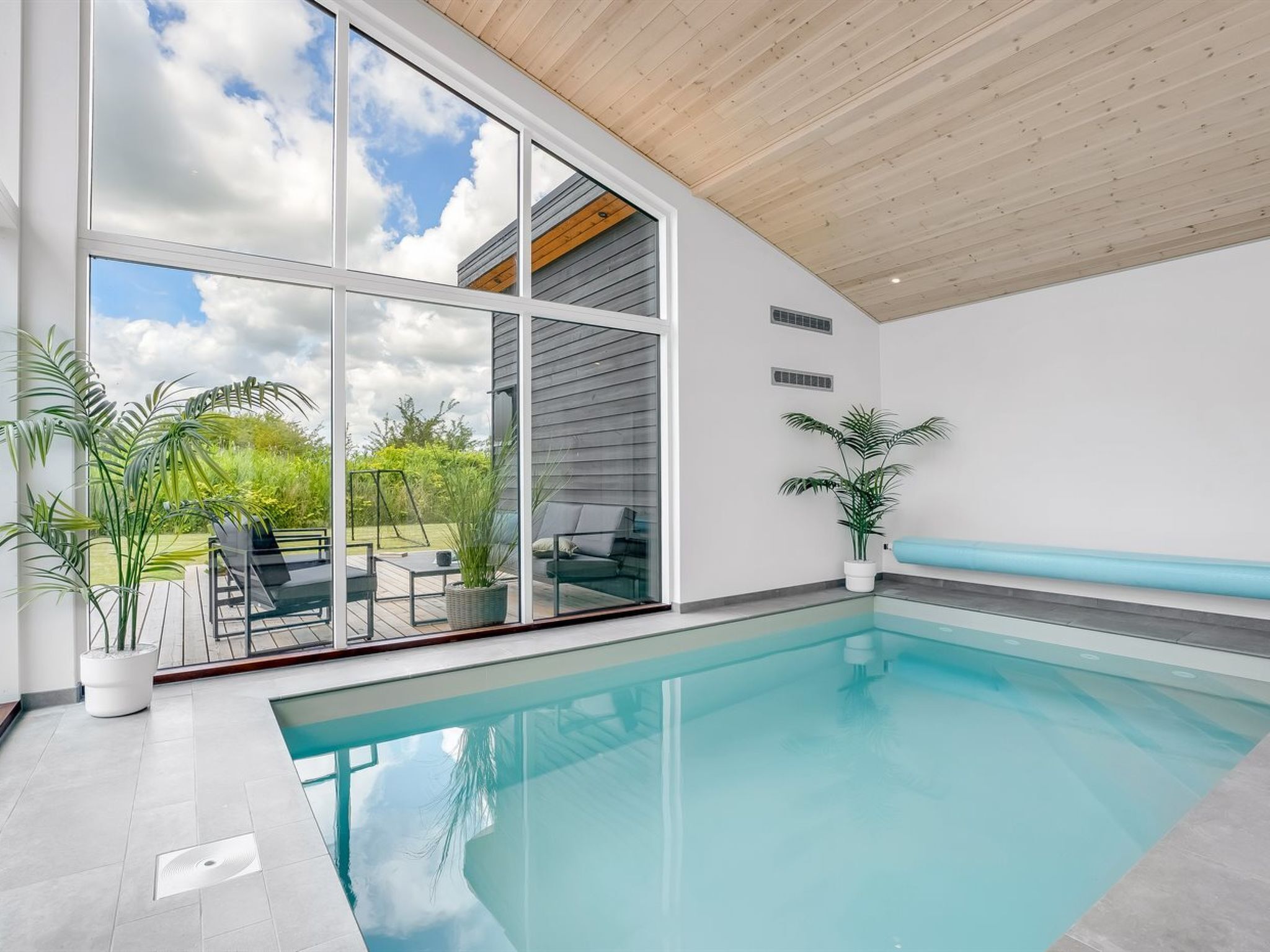 A modern indoor swimming pool with a glass wall, wooden ceiling, and a view of greenery outside.